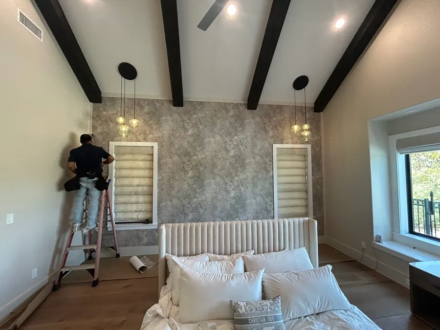 Craftsperson installing wallcovering in a vaulted bedroom with dark ceiling beams and pendant lighting.
